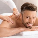 Happy middle aged man attending spa salon on weekend, closeup. Handsome bearded man smiling at camera while getting relaxing back massage. Female therapist rubbing joyful man shoulders at spa salon