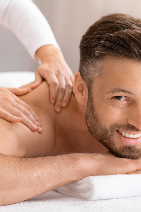Happy middle aged man attending spa salon on weekend, closeup. Handsome bearded man smiling at camera while getting relaxing back massage. Female therapist rubbing joyful man shoulders at spa salon