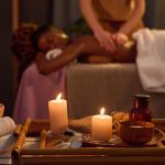 Medium close up of two lit aroma candles, jar full of essential oil on wooden tray and African American client lying on massage table and enjoying massaging